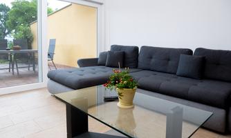 A modern living room with a gray sofa and a glass table. Through large windows, an outdoor area with a table and chairs is visible.