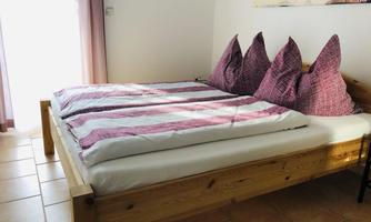 A cozy bedroom with a wooden bed and tall, upholstered pillows. The rooms are bright and friendly, with natural light streaming through the window.