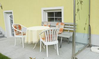 A cozy outdoor area with a round table and chairs. The wall is painted light yellow, and there is a small window front.