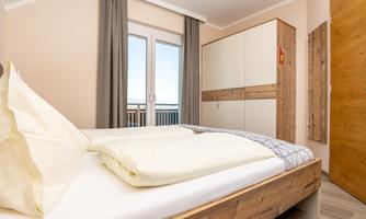 A modern bedroom with a double bed and light wood interior. Large windows provide plenty of daylight.
