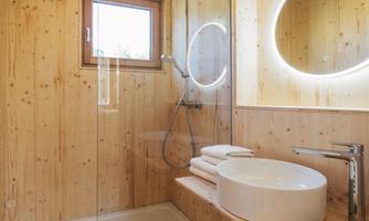 A modern bathroom with wooden panels and soft lighting. The shower is separated by a glass wall and a round mirror illuminates the space.