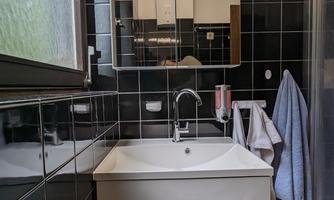 A modern bathroom with a white sink and a large mirror. The walls are in dark tiles, and there is a shower in the corner.