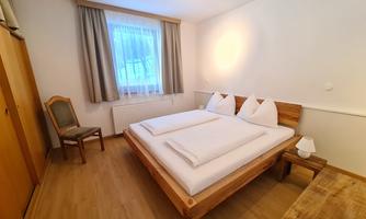 A simple bedroom with a wooden bed and white bedding. The room has a bright atmosphere with a window and minimalist furniture.