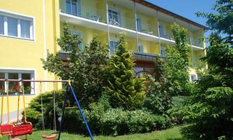 A yellow building with balconies and a well-maintained garden. In the foreground, there is a swing between the trees.