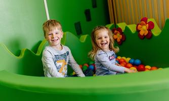 Two smiling children are sitting in a colorful ball pit. The surroundings are green with cheerful flowers.