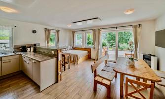A bright and modern living room with an open kitchen. Large windows provide a view of the green surroundings.