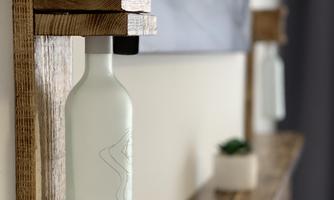 A modern wooden bottle holder mounted on a wall. The holder features a decorative, transparent bottle and is situated above a wooden shelf.