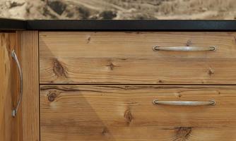 A modern kitchen drawer made of light wood with clear handles. The background features a gentle, natural landscape.