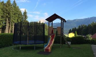 A playground with a slide and a trampoline on a green lawn. In the background, there are trees and mountains.