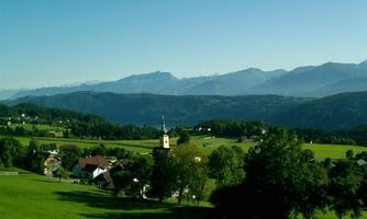 A picturesque landscape with gentle hills and trees. In the background, the mountains rise under a clear blue sky.