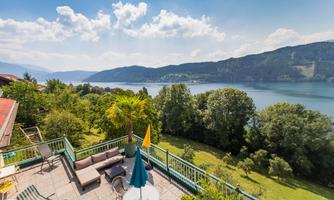 A terrace with cozy furniture and sun umbrellas, surrounded by trees and overlooking a picturesque lake. The weather is sunny and the landscape is green.
