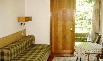 A simple room with a checkered sofa and a wooden chair. The door opens to a balcony overlooking nature.