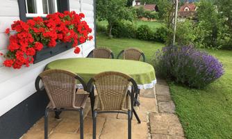 A beautiful outdoor area with a table and four chairs. In the background, red geraniums and lavender bloom next to a green meadow.