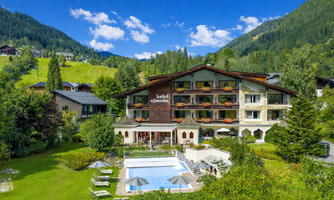 A picturesque hotel surrounded by mountains and green meadows. In the foreground, a swimming pool can be seen, surrounded by sun loungers.