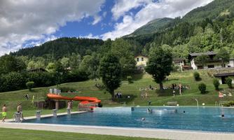 A swimming pool with a slide, surrounded by green meadows and trees. In the background are mountains and some people relaxing or swimming.