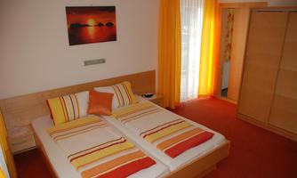 A cozy room with two single beds, colorful cushions, and a painting on the wall. The large windows let in a lot of light, and the curtains are in warm yellow tones.