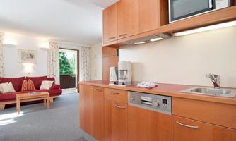 A modern kitchen with wooden furniture and a microwave. In the background, there is a cozy living area with a sofa and a window that leads to a balcony.