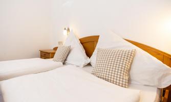 A bright bedroom with two single beds and checked pillows. The walls are white and there is a bedside lamp next to the bed.