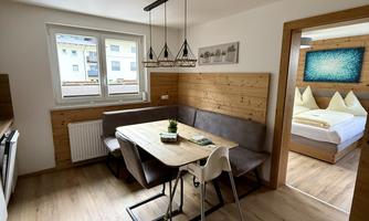 A cozy dining area with a wooden table and modern chairs. In the background, there is a bedroom featuring a large bed and decorative pillows.