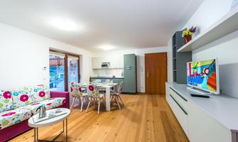 A modern living room with a colorful sofa and wooden floor. The open kitchen and dining area are well lit, and a television is visible.