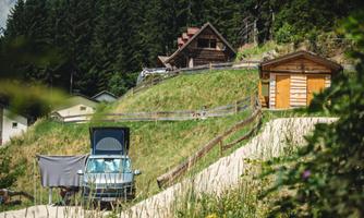 A motorhome is parked in a meadow with a view of a picturesque valley. In the background, several cabins and tall trees can be seen.