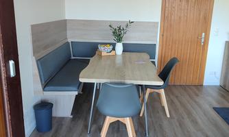 A modern furnished dining area with a wooden table and chairs. The walls are light, and there is a small bouquet of flowers on the table.