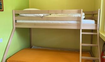 A room with a wooden bunk bed and an orange bed sheet on the lower mattress. The walls are painted light green, and there is a small chair next to it.