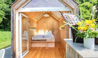 A cozy little wooden house with a bright interior and a double bed. In front of the house, there is a bouquet of flowers on a table, offering a view of the greenery.