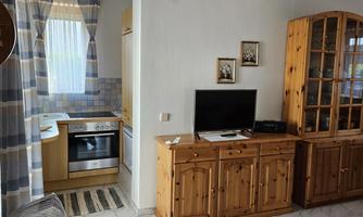 A cozy living area with a television and a wooden piece of furniture. In the background, a bright kitchen with a window can be seen.