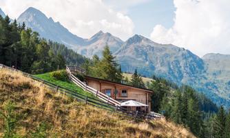 A cozy cabin on a hill, surrounded by green forests and tall mountains. The sky is clear with a few clouds.