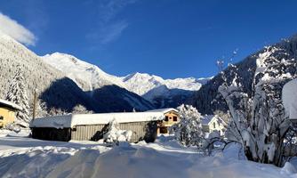 A snowy landscape with high mountains and clear blue sky. In the foreground, a few houses and snow-covered trees can be seen.