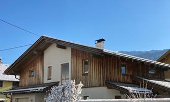 A beautiful wooden house with a snow-covered roof. The clear blue sky and the surrounding mountains create an idyllic winter landscape.