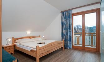 A cozy bedroom with a wooden bed and a window that lets in natural light. The view shows a balcony with a beautiful surrounding.