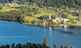 A picturesque landscape with a tranquil lake and gentle hills. Small houses and a church tower can be seen by the shore.