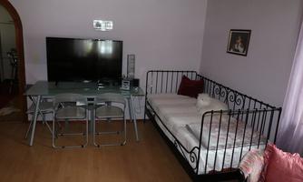 A cozy room with a bed, a glass table, and a television. The walls are painted in soft colors, and there is a small seating area.