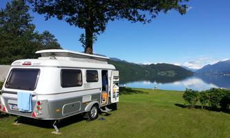 A caravan stands in a green meadow with a view of a tranquil lake. In the background, mountains and a blue sky can be seen.