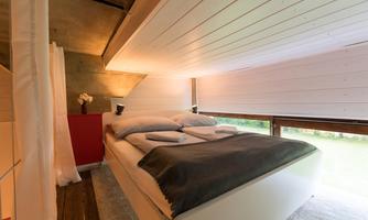 A modern bedroom with a cozy double bed and wood paneling. Large windows let in plenty of daylight and offer a view of the outdoors.