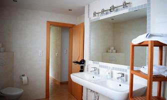 A modern bathroom with a double sink and large mirror. In the background, a toilet and a wooden shelf with towels can be seen.