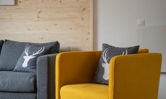 A cozy living area with a gray sofa and a bright yellow armchair. The cushions in the armchair feature a deer motif and the walls are made of light wood.