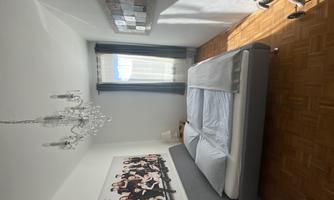 A cozy bedroom with a large bed and a crystal chandelier. Bright curtains let in light, and a decorative picture adorns the wall.