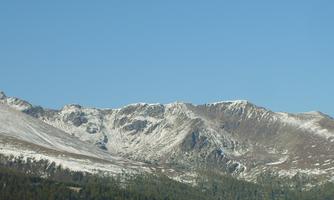 A mountain range with snow-covered peaks and clear blue sky. In the foreground, green forests can be seen.