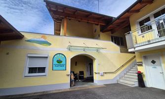 A yellow building with an entrance area and a staircase. The sign reading "FLATTACH" and a painted symbol depict a sunny landscape.