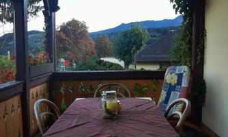 A cozy balcony with a table and chairs. In the background, trees and mountains can be seen, while the sky is blue.