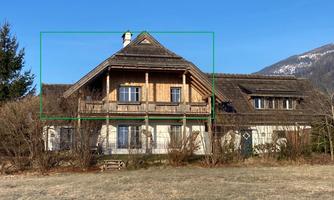 A large, traditional house with a wooden facade and balcony. In the background, mountains and a clear blue sky are visible.