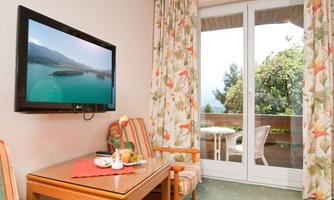 A cozy hotel room with a television, a table, and two chairs. The windows offer a view of a green oasis and a balcony.
