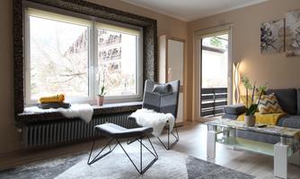 A stylishly furnished living room with a large window and a cozy armchair. Decorative plants are on the table and a few towels are neatly placed on the windowsill.