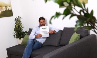 A man is sitting on a gray couch and holding a piece of paper in his hand. In the background, plants and pictures on the wall are visible.