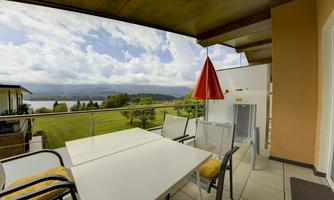A balcony with a table and chairs, surrounded by green meadows and a view of the lake. The sky is cloudy, but there are also bright spots.