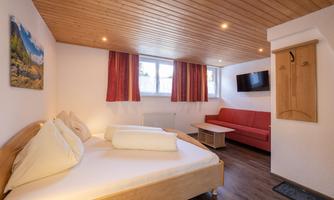 A bright room with wooden floor and ceiling. There is a double bed, a red couch, and a television on the wall.