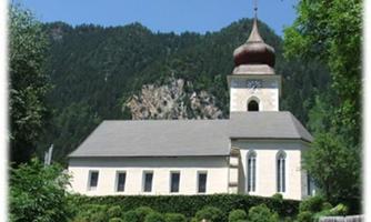 Eine malerische Kirche mit einem hohen Turm und einer schönen Fassade. Im Hintergrund sind grüne Berge und Bäume zu sehen.